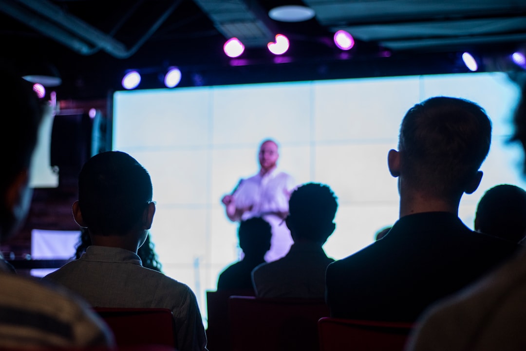 An audience watching a speaker on a bright screen in a dark room